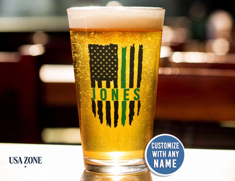 May include: A clear glass pint glass filled with golden beer, featuring a black and green distressed American flag graphic with the name JONES printed in green across the center. A blue circular badge reads CUSTOMIZE WITH ANY NAME.