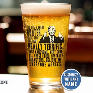 May include: A clear glass pint glass filled with golden beer, featuring a black graphic of a man giving two thumbs up and the text: YOU ARE A GREAT HUNTER. REALLY GREAT. THE BEST. REALLY TERRIFIC. VERY HANDSOME. VERY COOL. ALL THOSE OTHER HUNTERS? DISASTERS. BELIEVE ME. EVERYONE AGREES.