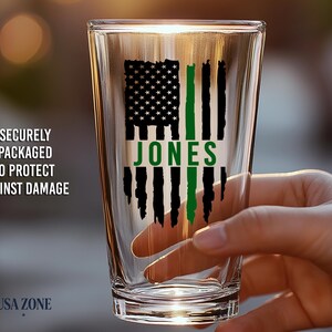 May include: A clear glass pint tumbler featuring a black and green graphic of a distressed American flag with the name JONES printed in green across the center. The design is printed on the front of the glass.