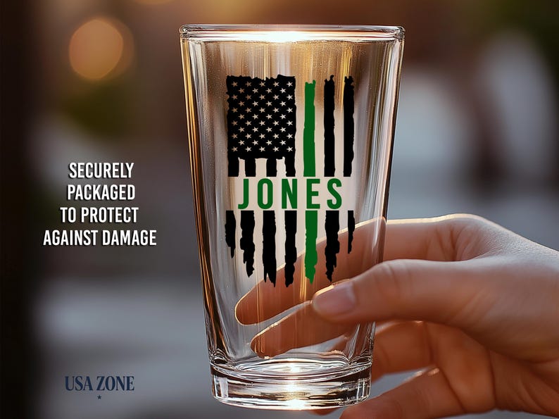 May include: A clear glass pint tumbler featuring a black and green graphic of a distressed American flag with the name JONES printed in green across the center. The design is printed on the front of the glass.