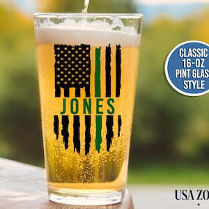 May include: A clear 16-oz pint glass filled with golden beer, featuring a black and green distressed American flag graphic with the name JONES printed in green across the center. A circular badge reads CLASSIC 16-OZ PINT GLASS STYLE.