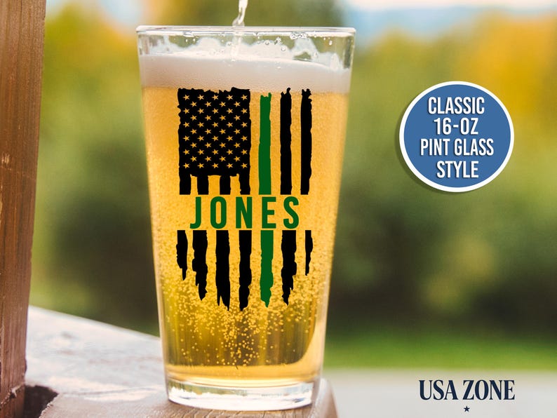 May include: A clear 16-oz pint glass filled with golden beer, featuring a black and green distressed American flag graphic with the name JONES printed in green across the center. A circular badge reads CLASSIC 16-OZ PINT GLASS STYLE.
