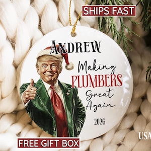 May include: A round white ceramic ornament featuring a graphic of a man in a green suit, red tie, and Santa hat giving a thumbs up. The text reads Andrew, Making PLUMBERS Great Again, and 2026. It hangs from a gold string.