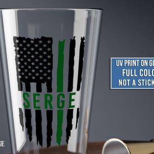 May include: A clear glass pint glass featuring a black and green distressed American flag graphic with the name SERGE printed in green. A blue rectangular box contains the text UV PRINT ON GLASS FULL COLOR NOT A STICKER.