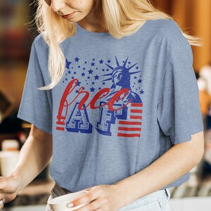 Peut inclure: Un t-shirt bleu clair avec un motif graphique rouge, blanc et bleu repr&eacute;sentant la Statue de la Libert&eacute;, des &eacute;toiles et des rayures. Le texte "free AF" est imprim&eacute; sur le t-shirt.