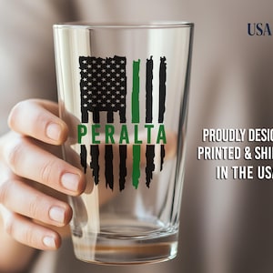 May include: A clear glass pint glass featuring a black and green distressed American flag graphic with the name PERALTA printed in green across the center. The design is printed on the front of the glass.