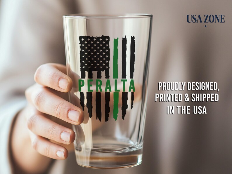 May include: A clear glass pint glass featuring a black and green distressed American flag graphic with the name PERALTA printed in green across the center. The design is printed on the front of the glass.