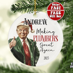 May include: A white ceramic Christmas ornament featuring a portrait of a man in a Santa hat and a green jacket, giving a thumbs-up. The ornament has the text "Andrew Making Plumbers Great Again 2025" in black and red.