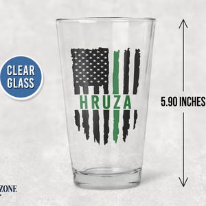May include: A clear glass pint glass measuring 5.90 inches in height. The glass features a black and green distressed American flag graphic with the name HRUZA printed in green across the center of the flag design.