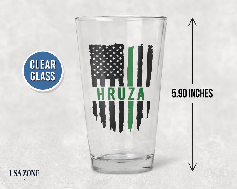 May include: A clear glass pint glass measuring 5.90 inches in height. The glass features a black and green distressed American flag graphic with the name HRUZA printed in green across the center of the flag design.