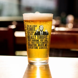 May include: A clear glass beer mug with a black and white graphic of a man with a thumbs up gesture. The text on the mug reads "You are a great plumber. Really great. The best. Really terrific. Very handsome. Very cool. All those other plumbers? Disasters. Believe me. Everyone agrees."