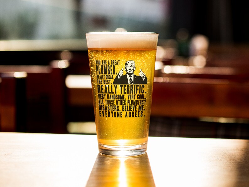 May include: A clear glass beer mug with a black and white graphic of a man with a thumbs up gesture. The text on the mug reads "You are a great plumber. Really great. The best. Really terrific. Very handsome. Very cool. All those other plumbers? Disasters. Believe me. Everyone agrees."