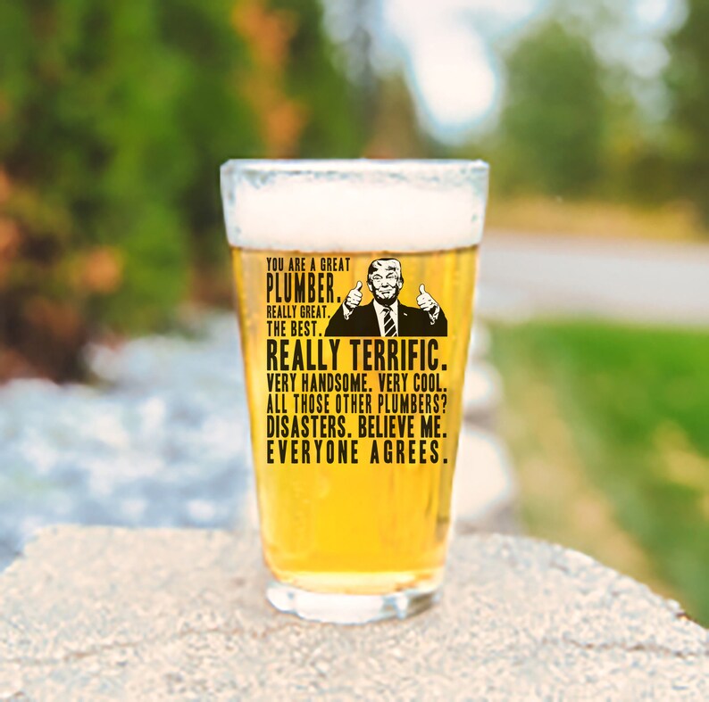 May include: A clear glass beer stein with a humorous graphic of a man with a thumbs up gesture and the text "You are a great plumber. Really great. The best. Really terrific. Very handsome. Very cool. All those other plumbers? Disasters. Believe me. Everyone agrees."