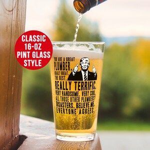 May include: A 16-ounce pint glass with a black and white image of a man with a thumbs up gesture. The text on the glass reads "You are a great plumber. Really great. The best. Really terrific. Very handsome. Very cool. All those other plumbers? Disasters. Believe me. Everyone agrees."