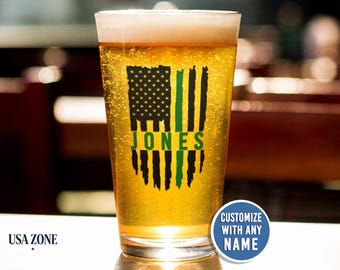 May include: A clear glass pint glass filled with golden beer, featuring a black and green distressed American flag graphic with the name JONES printed in green across the center. A blue circular badge reads CUSTOMIZE WITH ANY NAME.