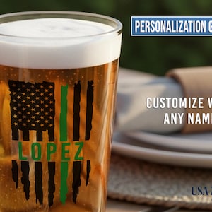 May include: A clear glass pint beer mug filled with amber liquid and white foam. The glass features a black distressed American flag graphic with a vertical green stripe and the name LOPEZ in green text. Text reads PERSONALIZATION GUIDE.