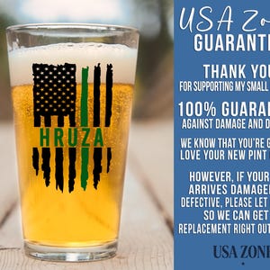 May include: A clear pint glass filled with golden beer, featuring a black and green graphic of a distressed American flag with the name HRUZA printed in green. The text USA ZONE GUARANTEE and additional business information appear to the right.