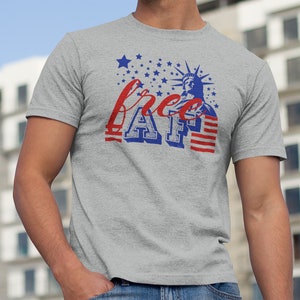 Peut inclure: Un t-shirt gris avec un motif d&eacute;lav&eacute; de la Statue de la Libert&eacute; et le texte "free AF" en bleu et rouge. Le motif est entour&eacute; d'&eacute;toiles blanches sur un fond bleu.