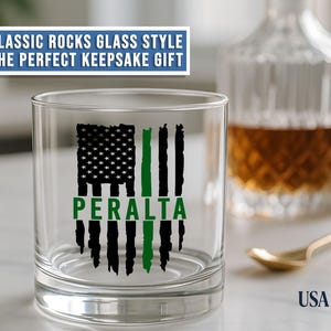 May include: A clear rocks glass featuring a black and green distressed American flag graphic with the name PERALTA printed in green across the center. The text CLASSIC ROCKS GLASS STYLE THE PERFECT KEEPSAKE GIFT appears above the glass.