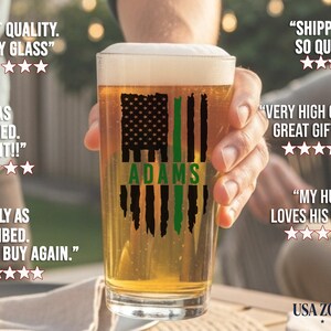 May include: A clear glass pint beer glass filled with golden beer and white foam. The glass features a black and green distressed American flag graphic with the name ADAMS printed in green across the center of the flag design.