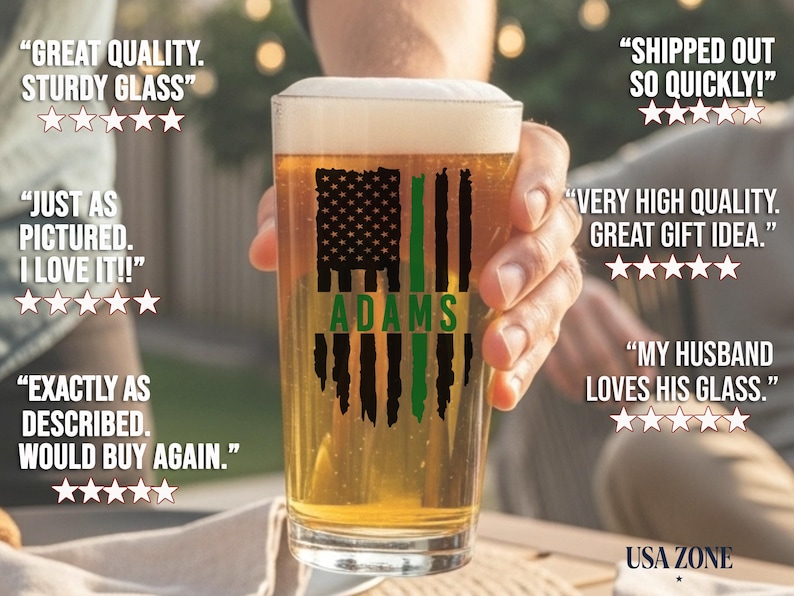 May include: A clear glass pint beer glass filled with golden beer and white foam. The glass features a black and green distressed American flag graphic with the name ADAMS printed in green across the center of the flag design.
