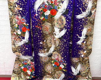 May include: A vibrant purple kimono featuring white cranes in flight, floral arrangements, and gold accents. The traditional Japanese garment has a white, fluffy hem and is displayed against a white brick wall.