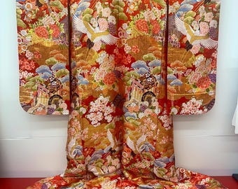 May include: A vibrant, traditional Japanese kimono with a red base and intricate gold detailing. The design features white cranes, flowers, and clouds. The kimono is displayed on a hanger, showcasing its full length and ornate patterns.