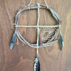 May include: A handmade dreamcatcher with a wooden hoop, brown leather, and feathers. The dreamcatcher is decorated with a white and brown web and a turquoise bead.