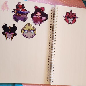 Op de afbeelding: Een open notitieboek met een witte kaft en een spiraalbinding. Er zijn verschillende kleurrijke stickers op de pagina's, waaronder een cartoonrobot, een meisje in een rode jurk en een zwart-wit wezen.