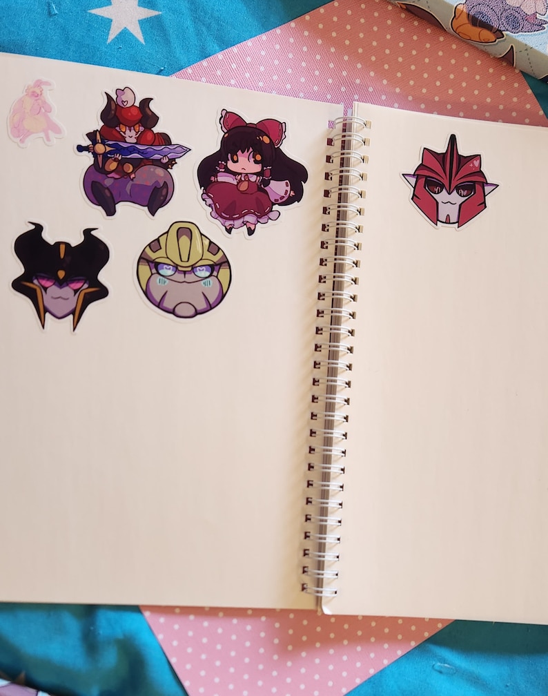 Op de afbeelding: Een open notitieboek met een witte kaft en een spiraalbinding. Er zijn verschillende kleurrijke stickers op de pagina's, waaronder een cartoonrobot, een meisje in een rode jurk en een zwart-wit wezen.