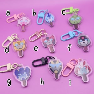 May include: Nine different keychains featuring cartoon characters from the Transformers franchise. Each keychain is a clear plastic charm shaped like an ice cream pop with a different character on each. The characters are all in different colours and poses. The keychains are attached to metal rings.