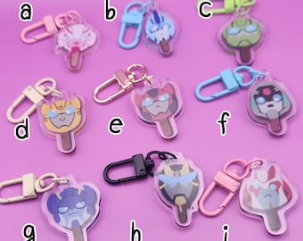 May include: Nine different keychains featuring cartoon characters from the Transformers franchise. Each keychain is a clear plastic charm shaped like an ice cream pop with a different character on each. The characters are all in different colors and poses. The keychains are attached to metal rings.