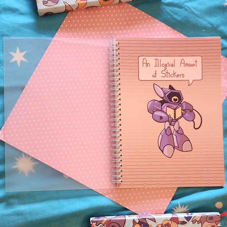 Op de afbeelding: Een roze spiraalnotitieblok met een wit stippenpatroon. De omslag toont een cartoonrobot die een boek vasthoudt en een spraakballon met de tekst "Een onlogische hoeveelheid stickers".