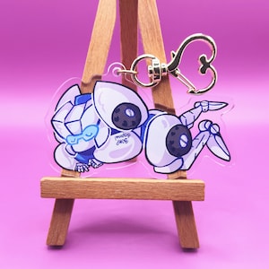 May include: A clear acrylic charm depicting a cartoon robot in white and blue with heart-shaped details. The charm has a gold-coloured clasp and the text "BOMP disposal". The charm is displayed on a small wooden easel against a pink background.