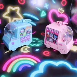 May include: Two small plastic van-shaped keychains in light blue and light pink. The blue van features a graphic of robots and the text DOWN WITH MEGATRON. The pink van features a graphic of robots and the text AUTOBUS. Both have clear windows.