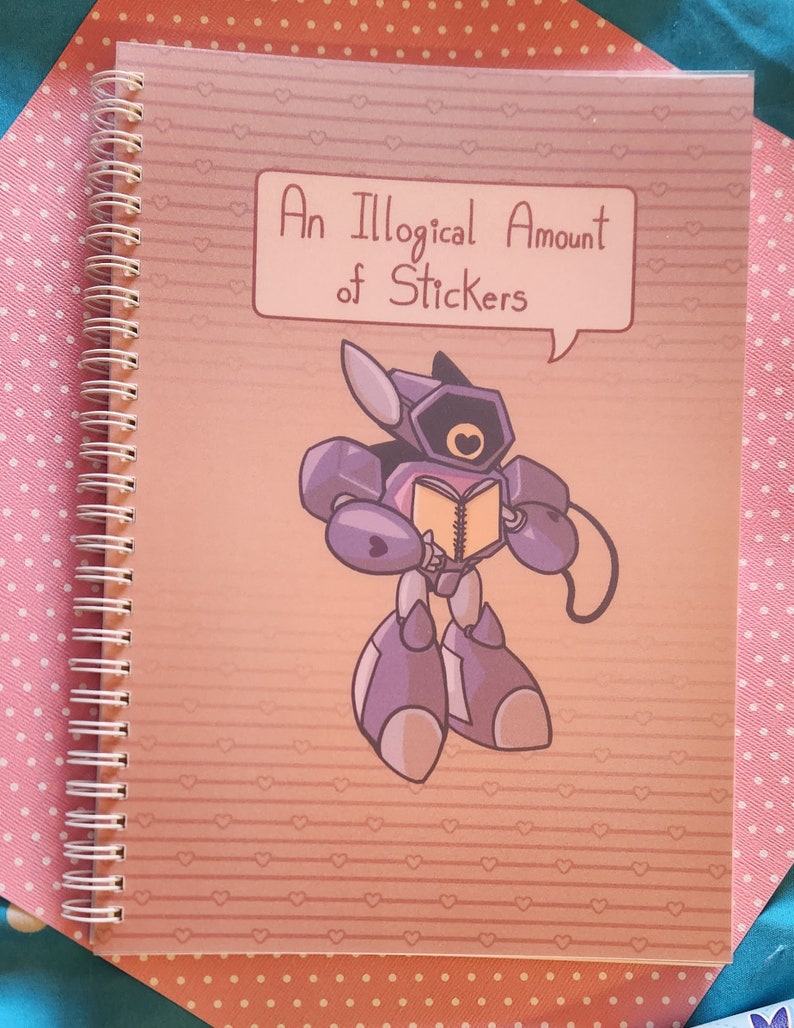 Op de afbeelding: Een roze spiraalnotitieblok met een roze en wit hartpatroon. De omslag heeft een cartoonrobot die een boek vasthoudt en een spraakballon met de tekst "Een onlogische hoeveelheid stickers".