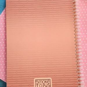 Op de afbeelding: Een roze spiraalgebonden notitieboek met een roze en wit hartpatroon op de kaft. Het notitieboek heeft een sticker op de voorkant met de tekst "Smoketopus".