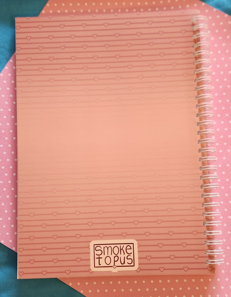 Op de afbeelding: Een roze spiraalgebonden notitieboek met een roze en wit hartpatroon op de kaft. Het notitieboek heeft een sticker op de voorkant met de tekst "Smoketopus".