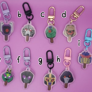 Op de afbeelding: Negen kleurrijke sleutelhangers met cartoonfiguren op een lollystok. De personages zijn uit de Transformers-franchise. De sleutelhangers zijn gemaakt van helder acryl en hebben een zilveren, gouden of zwarte metalen ring.