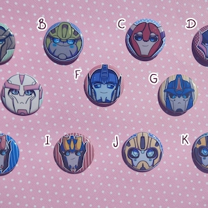 Op de afbeelding: Tien ronde, kleurrijke, cartoonfiguur-knoppen met Transformers-personages. De knoppen zijn gerangschikt in twee rijen van vijf. De personages zijn Optimus Prime, Bumblebee, Starscream, Arcee, Bulkhead, Ironhide, Wheeljack, Cliffjumper, Sideswipe en Jazz.