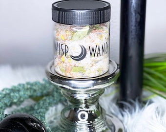 May include: A clear jar with a black lid, labeled "WISP WAND", sits on a silver pedestal. The jar contains colorful herbs and crystals. A tall black candle and a black sphere are also visible, along with a cluster of dark crystals.