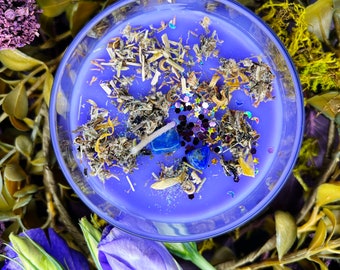 Op de afbeelding: Een ronde, heldere glazen kaars gevuld met lavendelkleurige was. De kaars is versierd met gedroogde kruiden, blauwe edelstenen en glitter. De kaars is omgeven door paars en groen gebladerte en blauwe bloemen.