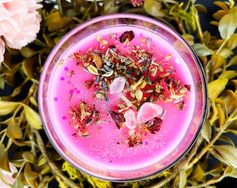 May include: A pink candle in a clear glass container, adorned with dried herbs, rose petals, and crystals. The candle is surrounded by greenery and a bundle of dried herbs wrapped in pink twine. A pink flower is visible in the background.