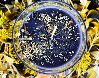 May include: A black candle in a clear glass container, adorned with dried herbs, glitter, and a white wick. The candle is surrounded by yellow-green foliage, creating a natural and mystical aesthetic. The candle's surface is speckled with colorful glitter.