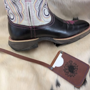 May include: A black and white leather cowboy boot with pink stitching. A brown leather card holder with a sunflower design is in the foreground. The card holder has a long strap and purple stitching.