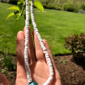 May include: White shell necklace with a turquoise turtle charm. The necklace is made of small, round shells strung together on a white cord.