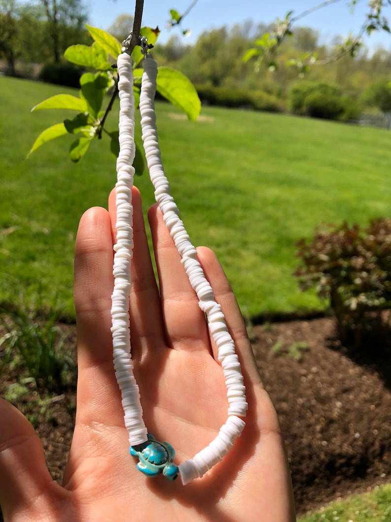 May include: White shell necklace with a turquoise turtle charm. The necklace is made of small, round shells strung together on a white cord.