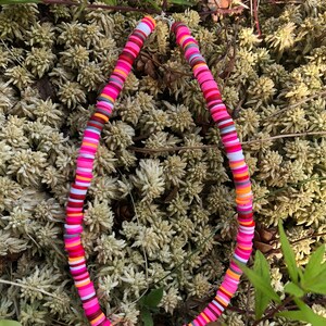 K&ouml;nnte beinhalten: Eine bunte Halskette aus runden, flachen Perlen in den Farben Pink, Orange, Wei&szlig; und Grau. Die Halskette liegt auf einem Bett aus gr&uuml;nem Moos.