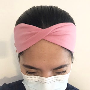 Puede incluir: Una diadema de tela rosa con un diseño de nudo retorcido. La diadema la lleva puesta una persona con el pelo oscuro y una mascarilla blanca.