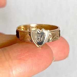 May include: A gold ring with a shield-shaped design featuring an engraved monogram. The ring band is decorated with a textured pattern.